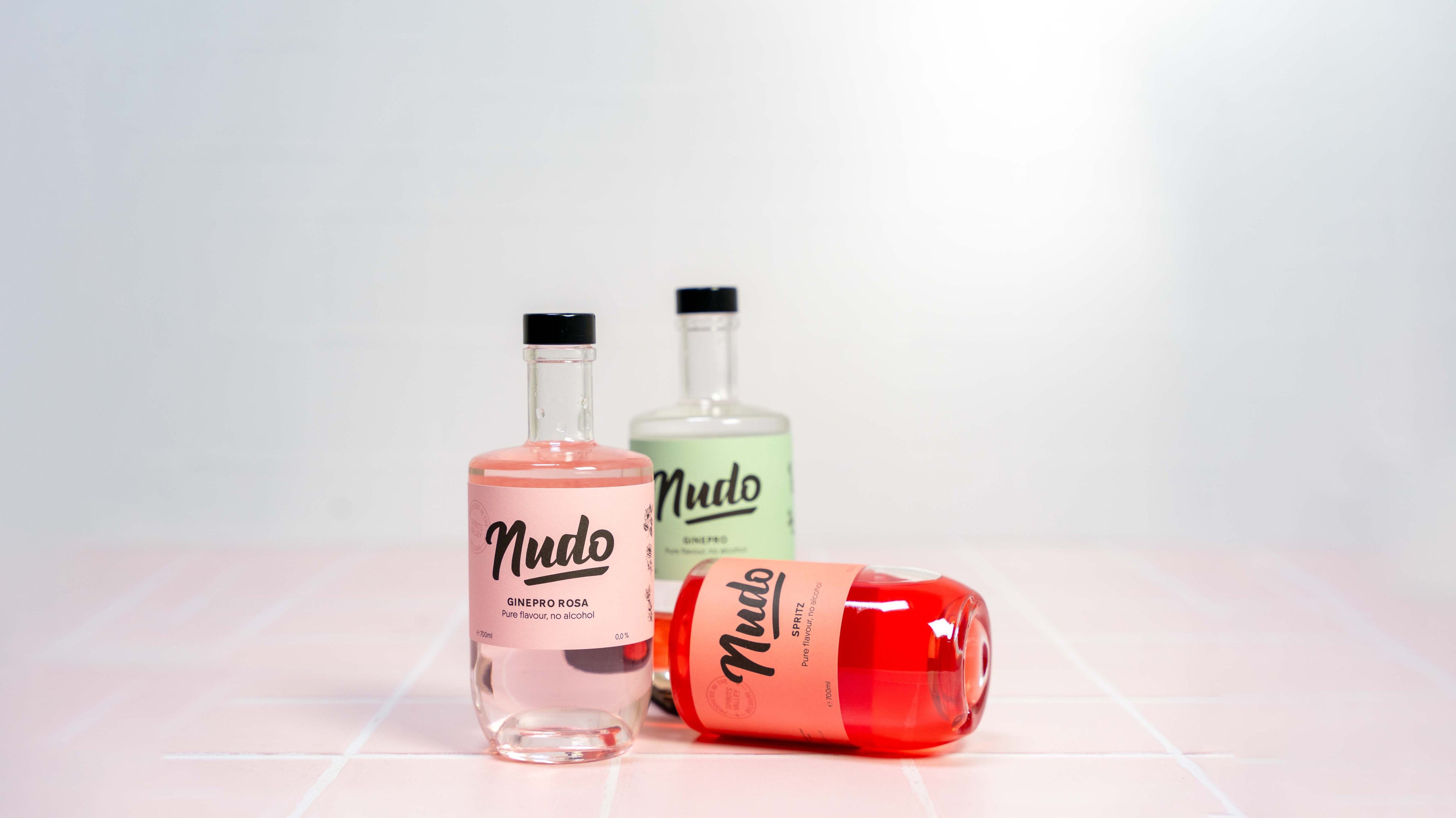 Drie flessen NUDO non-alcoholische spirits: Ginepro Rosa, Ginepro, en Spritz, op een roze tegeloppervlak met een minimalistische achtergrond.