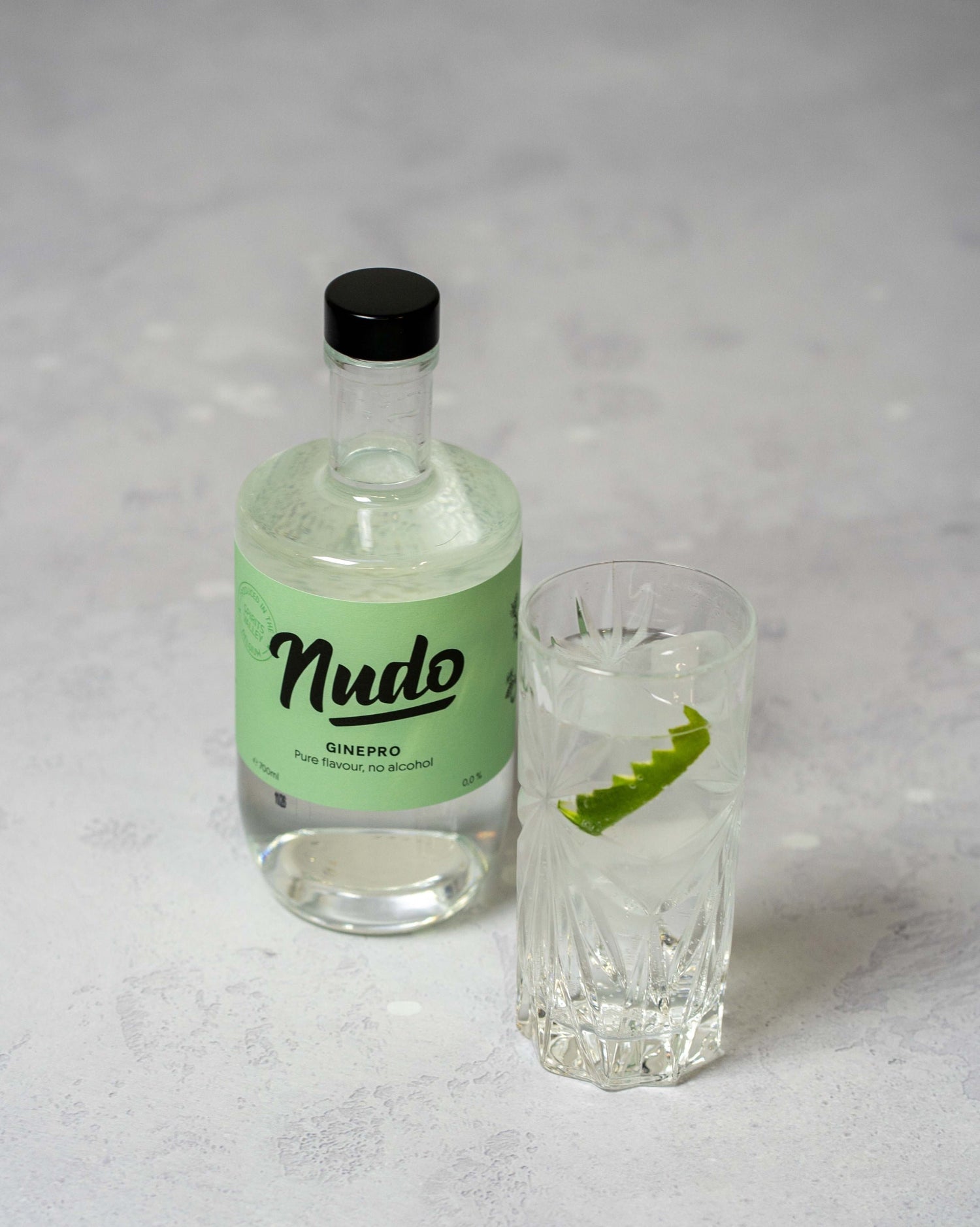 Fles NUDO Ginepro non-alcoholische drank op een lichtgrijze achtergrond, naast een glas gevuld met ijs en een garnering van limoen.