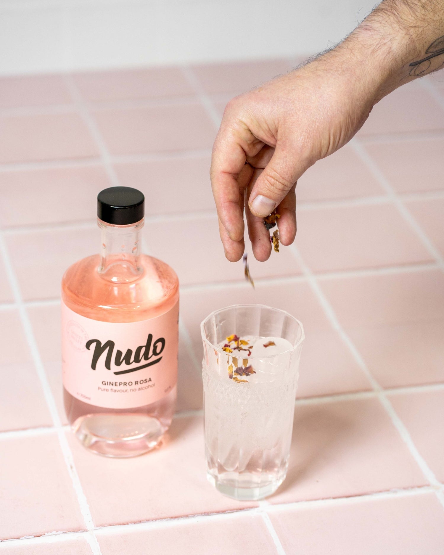 Fles NUDO Ginepro Rosa non-alcoholische drank op een roze tegeloppervlak, met een glas gevuld met ijs en een hand die rozenblaadjes toevoegt aan het drankje.