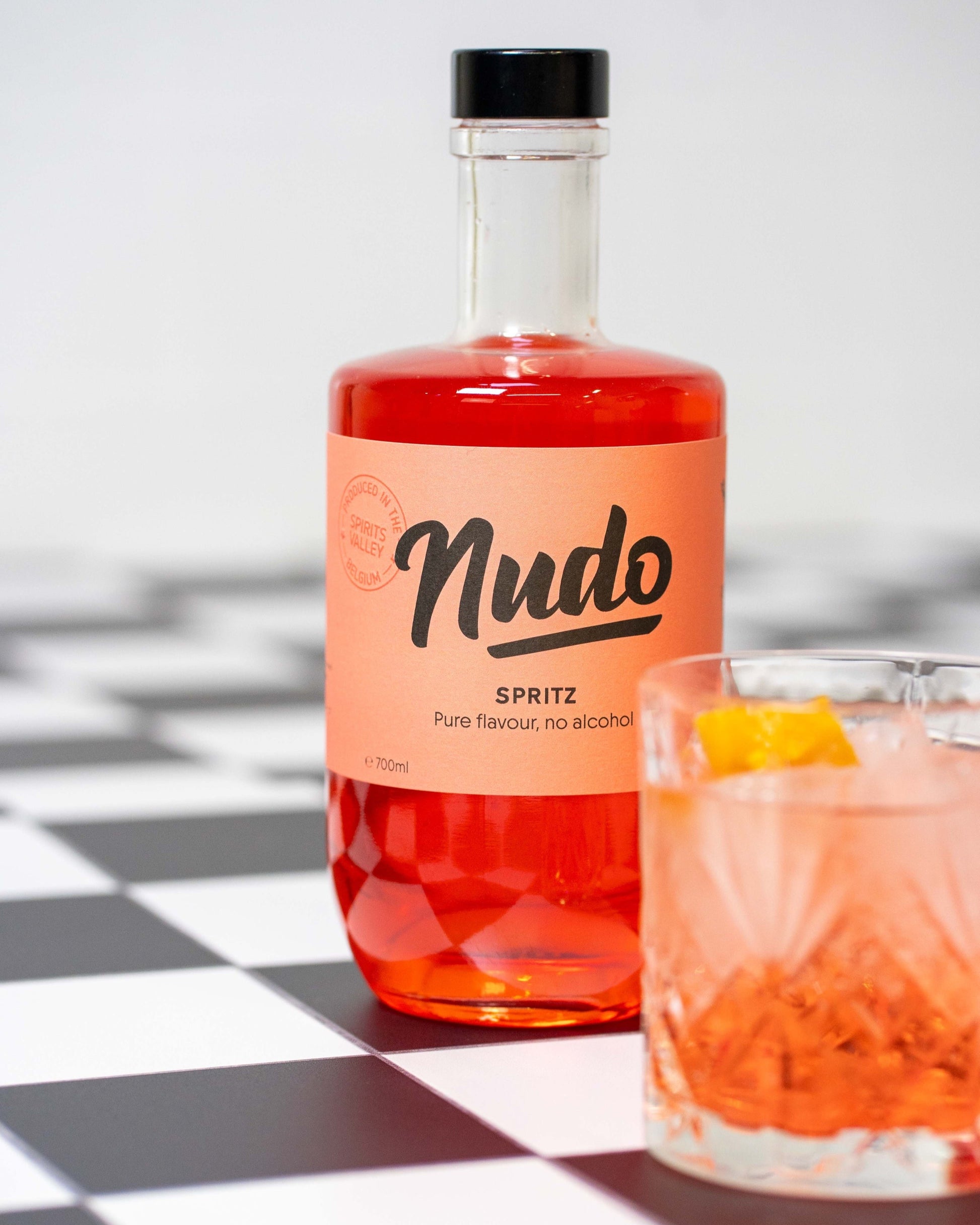 Fles NUDO Spritz non-alcoholische drank met een glas gevuld met ijs, NUDO Spritz en een schijfje sinaasappel op de voorgrond.