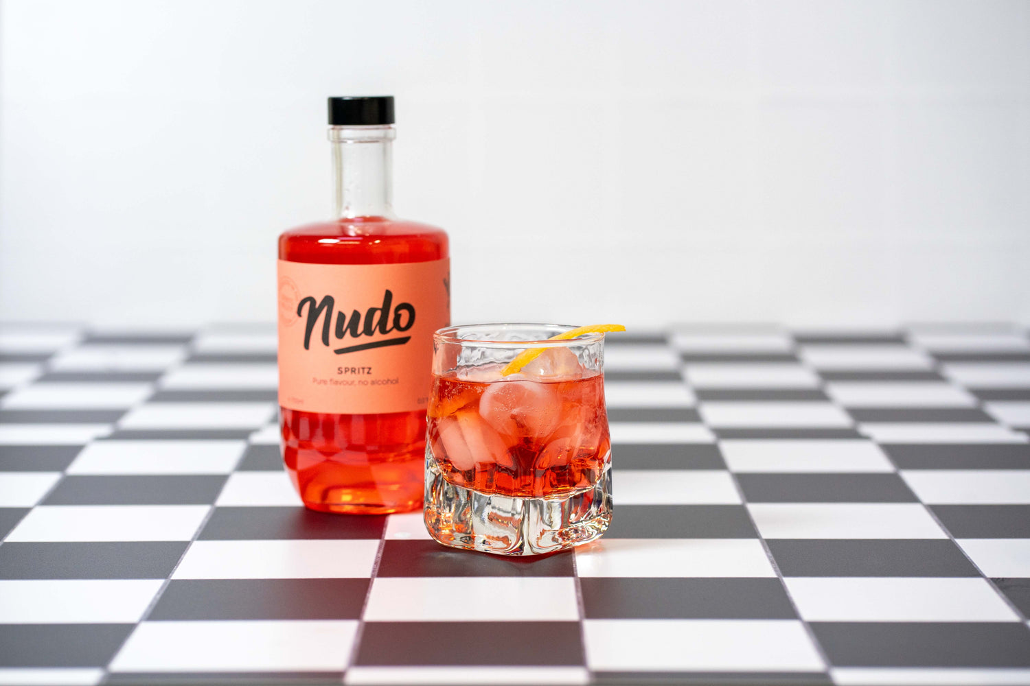 Fles NUDO Spritz non-alcoholische drank op een zwart-wit geblokt oppervlak, naast een glas gevuld met ijs, NUDO negroni en een schijfje sinaasappel als garnering.