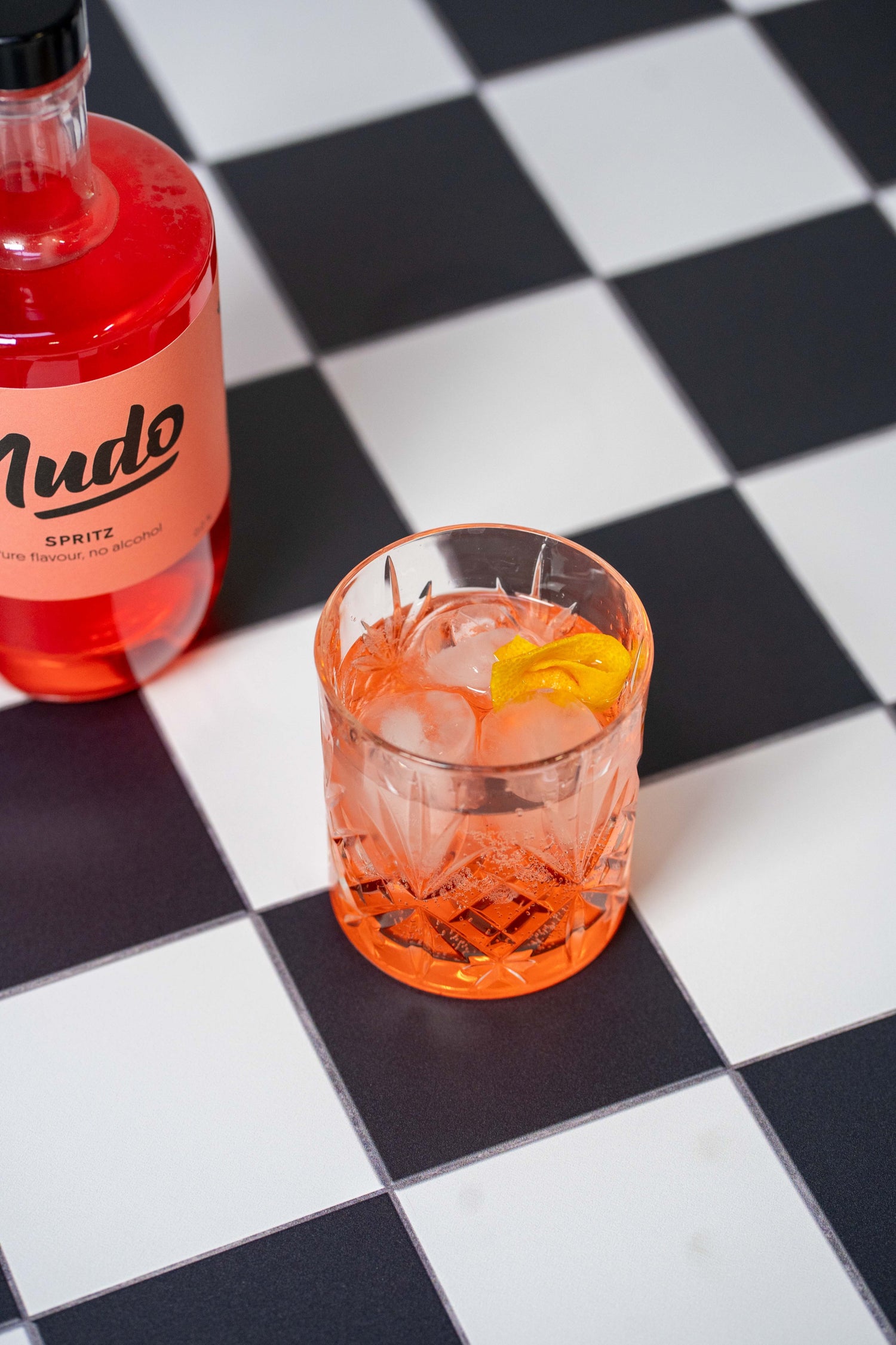 Close-up van een fles NUDO Spritz non-alcoholische drank op een zwart-wit geblokt oppervlak, naast een glas gevuld met ijs, Spritz en een garnituur van sinaasappelschil.