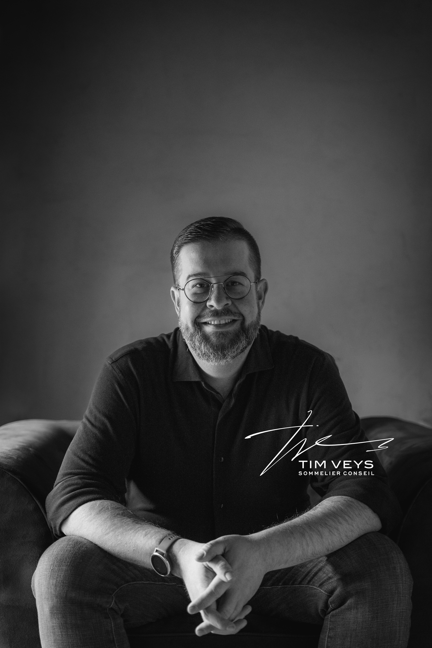 Zwart-wit portret van Tim Veys, Sommelier Conseil, zittend met gekruiste handen en een glimlach, met zijn handtekening en naam/logo in de rechterhoek.
