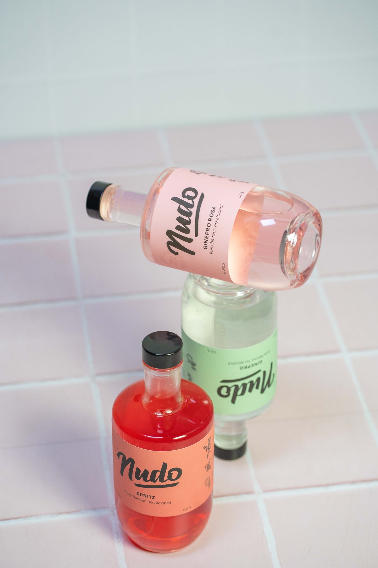 Drie flessen NUDO non-alcoholische spirits op een roze tegeloppervlak: Ginepro Rosa balancerend bovenop Ginepro, met Spritz op de voorgrond.